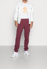 Vit denimjacka över en orange Adidas-logotyp t-shirt, tillsammans med vinröda sweatpants med sidofickor och vita sneakers.