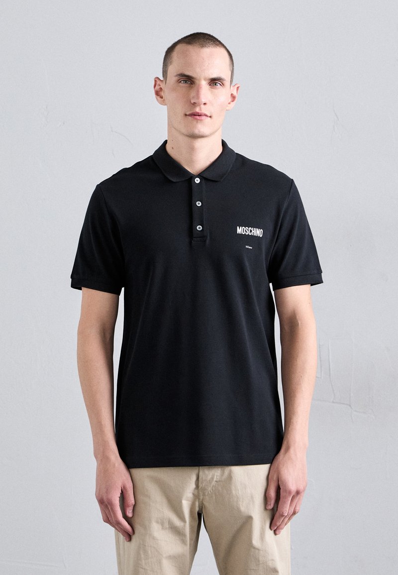 Juodas polo marškinėliai iš lygaus medvilnės, su klasikinis apykaklė, trijų mygtukų užsegimas ir baltas "Moschino" logotipas kairėje krūtinės pusėje.