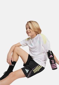 Weißes Sportshirt mit mehrfarbigen Streifen, schwarze Shorts mit Mustern, schwarze Wasserflasche mit pinken Akzenten und schwarze Socken.
