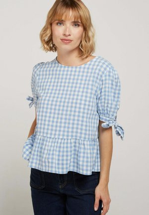 Femme portant un chemisier à carreaux bleu clair et blanc avec des manches courtes bouffantes nouées en nœuds, associée à un jean foncé, debout devant un arrière-plan uni.