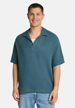 Homme souriant portant une chemise ample texturée à manches courtes sarcelle et un jean bleu clair, debout devant un fond clair uni.