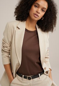 Beige blazer med struktureret stof, brun t-shirt og sort bælte med en cirkulær guldspænde. Krøllet hår og subtil makeup er synlig.