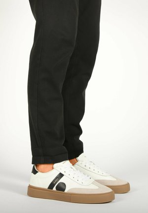 Sneakers bianche con accenti neri e beige, design con lacci, suole in gomma e stile minimalista, abbinate a pantaloni neri a gamba affusolata.
