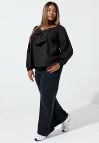 Svart blus med en stor rosettdetalj vid halsen, puffärmar, ihop med mörka utsvängda jeans och vita sneakers. Slät tygtextur.