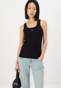 Svart ribbet tank topp med dyp utrigning og liten hvit logo på brystet, parret med lys blå denimjeans og en svart håndveske.