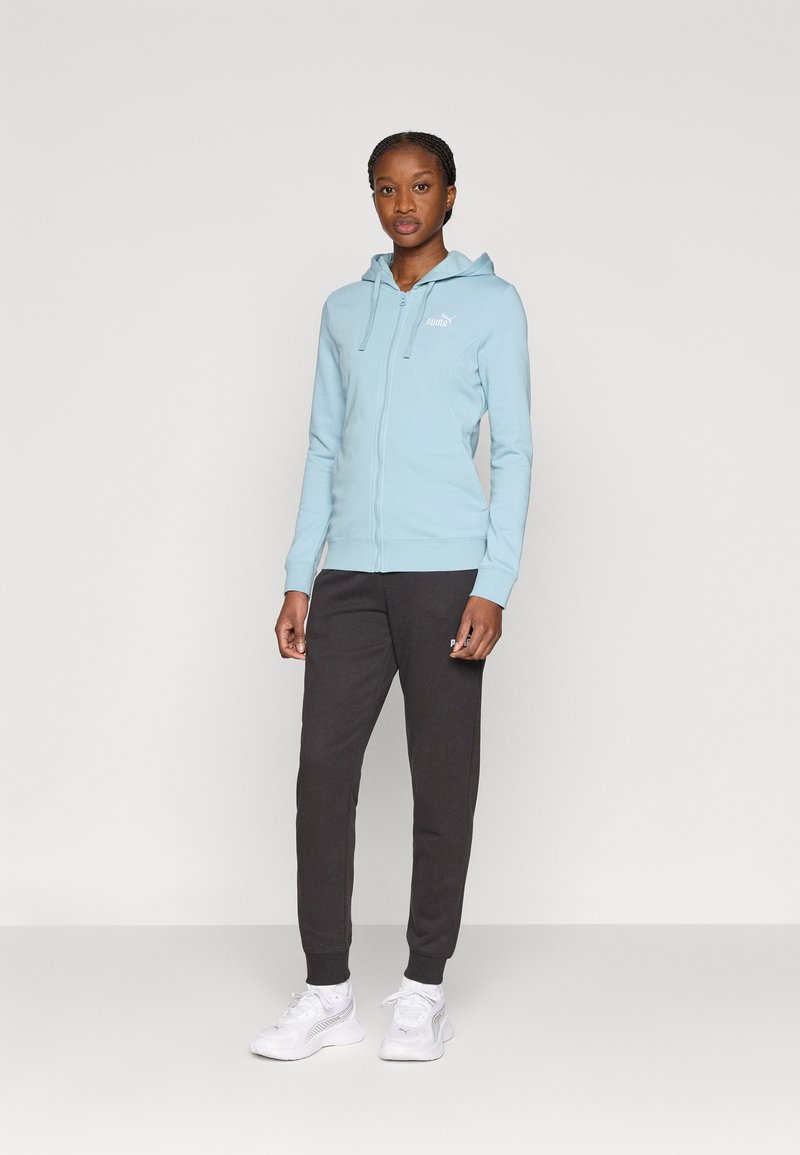 Ανοιχτό γαλάζιο φούτερ με φερμουάρ, κουκούλα και μπροστινές τσέπες, συνδυασμένο με σκούρο γκρι φόρμα και λευκά αθλητικά παπούτσια. Διαθέτει μικρό λογότυπο στο φούτερ.