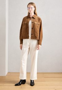Femme portant des lunettes bleues, une veste marron à boutons, un haut blanc, un pantalon en velours côtelé blanc et des bottes à talons noirs, debout sur un sol en bois.