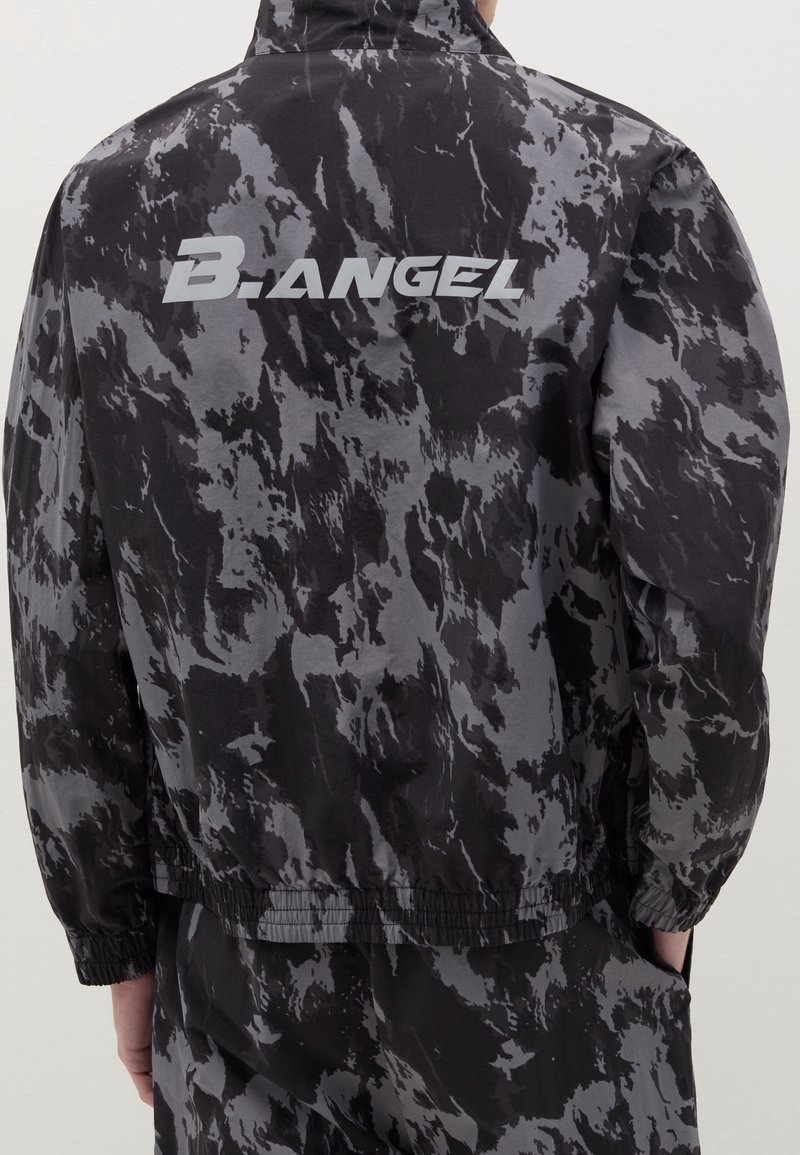 Personne portant une veste noire et grise à motif camouflage avec le texte "B.ANGEL" imprimé en blanc dans le dos.