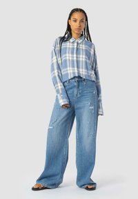Chemise surdimensionnée à carreaux bleu clair avec des manches longues et un col, associée à un jean en denim large et déchiré. Des sandales plates complètent le look.