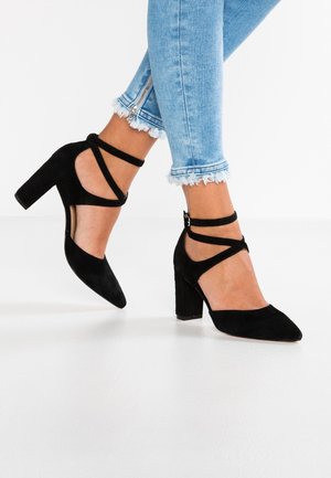 Kvinde iført sorte ruskind blokhæle med ankelremme parret med lyseblå skinny jeans med frynset kant og lynlåse.
