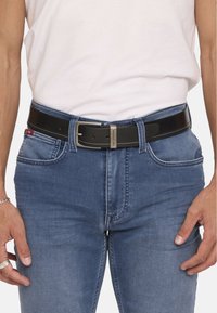 Zwarte leren riem met een gestructureerde afwerking, voorzien van een zilveren gesp en contrasterende stiksels, gedragen over blauwe spijkerbroek.