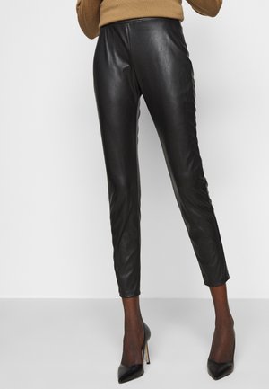 Smaltsiddende sorte bukser i kunstlæder båret med sorte gennemsigtige strømpebukser og sorte spidse højhælede sko, kombineret med en kamel-farvet sweater.