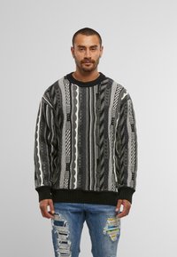 Schwarz-weißer, gemusterter Pullover mit strukturiertem Strick, Rundhalsausschnitt und gerippten Bündchen, getragen über abgetragenen blauen Jeans.