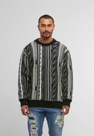 Schwarz-weißer, gemusterter Pullover mit strukturiertem Strick, Rundhalsausschnitt und gerippten Bündchen, getragen über abgetragenen blauen Jeans.