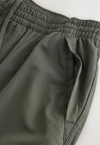 Rohelised spordipüksid, millel on elastne vöökoht, küljetaskud ning sile ja kergelt läikiv kangas.