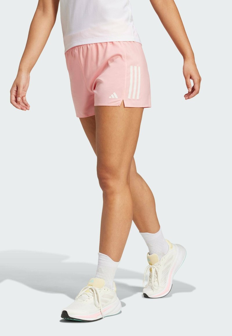 Helle pinke Sportshorts mit einem weißen Drei-Streifen-Design. Verfügt über einen elastischen Bund und seitliche Schlitze für mehr Bewegungsfreiheit. Kombiniert mit weißen Turnschuhen.