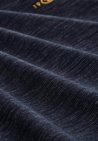 Námorný textil s hladkým, textúrovaným povrchom, s jemnými vlnkovými vzormi a svetlými bodkami, doplnený zlatým akcentovaným logom.