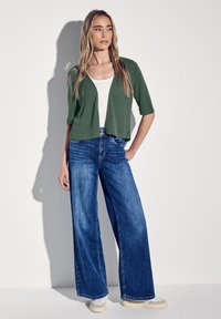 Cardigan court vert à manches courtes sur un haut blanc, associé à un jean bleu taille haute et large. Des chaussures légères complètent la tenue.