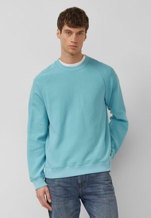 Młody mężczyzna z krótkimi brązowymi włosami, ubrany w jasnoniebieską bluzę i niebieskie dżinsy, stoi na tle jednolitego szarego tła.