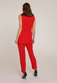 Röd ärmlös tunika ihop med matchande skräddarsydda byxor, gjorda av ett slätt tyg, med rak skärning och elegant silhuett. Svarta högklackade skor.