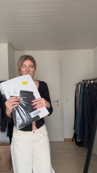 Zwarte leggings in plastic verpakking gehouden tegen een zwart shirt en crèmekleurige broek met wijde pijpen. Op de achtergrond kleding aan hangers.