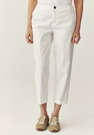 Osoba ubrana w białe, krótkie, dopasowane spodnie z przednim kantem oraz beżowe sneakersy na sznurowadła, stojąca na tle jednolitego tła.