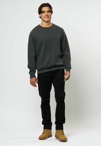 Szürke, hosszú ujjú pulóver, bordás mandzsettákkal, kerek nyakkivágással és egy kis logóval. Fekete farmernadrággal és bézs csizmával párosítva.