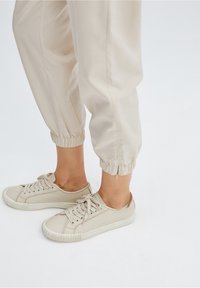 Beige elastische manchet joggingsbroek gecombineerd met lichtbeige sneakers. Sneakers hebben een ronde neus, vetersluiting en een gestructureerde rubberen zool.