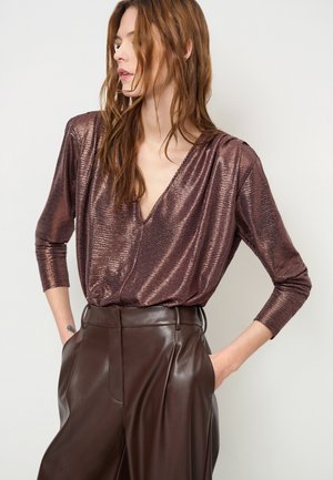 Vrouw draagt een glanzende, gestructureerde bronzen top met lange mouwen en hoog getailleerde bruine leren broek, met handen in de zakken, kijkt opzij.