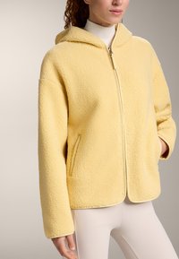 Jachetă galbenă din fleece, cu fermoar frontal și glugă. Are mâneci supradimensionate și buzunare laterale, cu o suprafață moale și texturată.