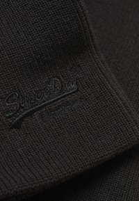 Tessuto nero a coste con un aspetto texturizzato, caratterizzato da un logo ricamato in evidenza con filo scuro. Nessun motivo o decorazione aggiuntiva.