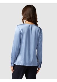 Light blue long-sleeve blouse in smooth, shiny fabric. Features a rounded neckline and buttoned cuffs. Falls to mid-hip length.