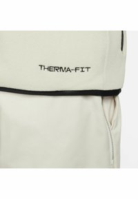 Chaqueta de forro polar en color blanco roto con detalles en negro. Presenta el logotipo bordado "THERMA-FIT" y un bolsillo lateral visible con cremallera. Textura suave.