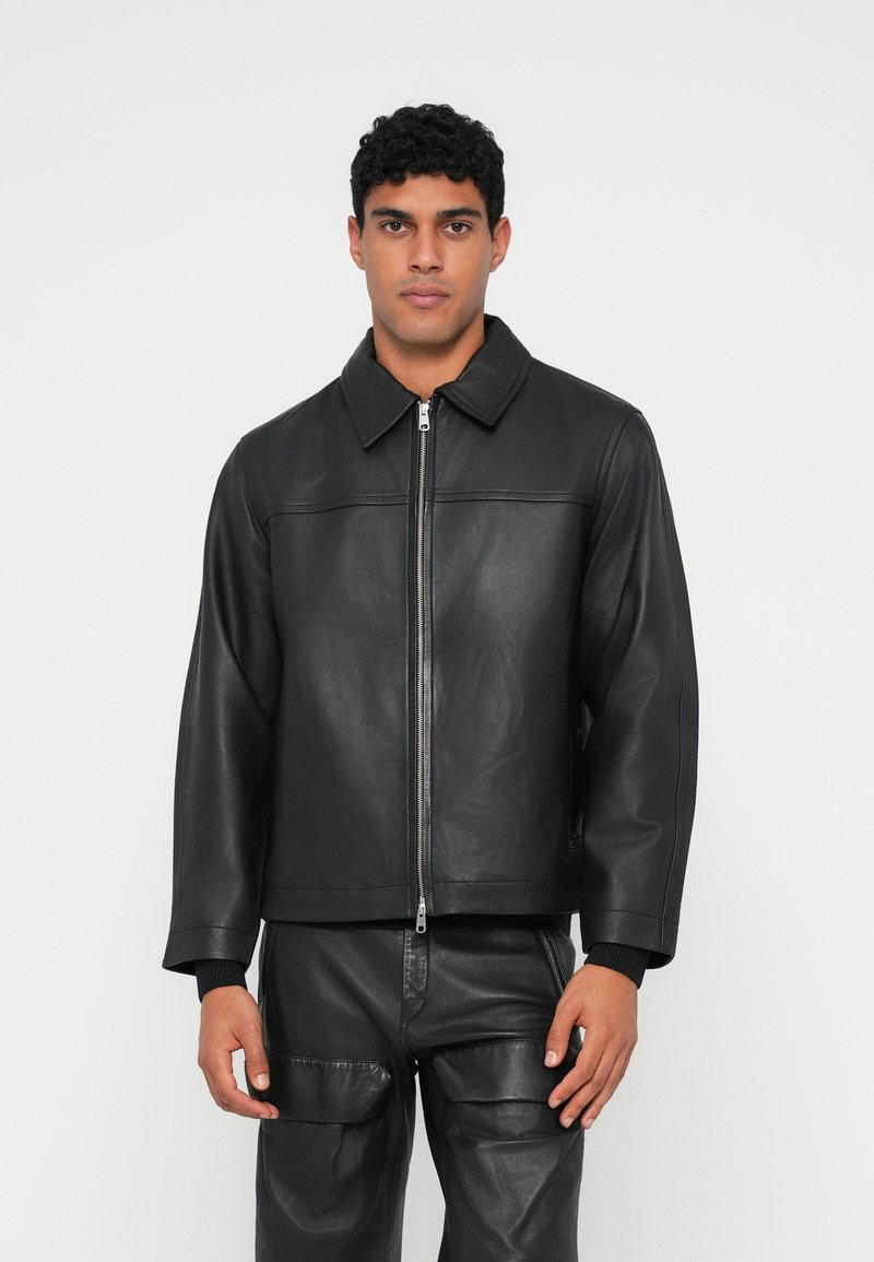 Chaqueta de cuero negro con cierre delantero, cuello ancho y puños acanalados. Textura suave con líneas limpias y un minimalismo en los detalles.