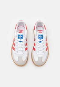 Adidas-sneakers i vit läder med röda ränder och en beige gummisula. Har en blå logotyp och snörningssystem på en rundad tåkonstruktion.