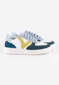 Une paire de baskets blanches avec des accents en daim teal et jaune, des lacets bleu clair et un onglet au talon bleu clair sur un fond blanc.