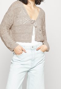 Cárdigan de punto beige con escote en V, frente abierto y patrones intrincados. Presenta mangas largas y un diseño cropped. Combinado con jeans de color azul claro.