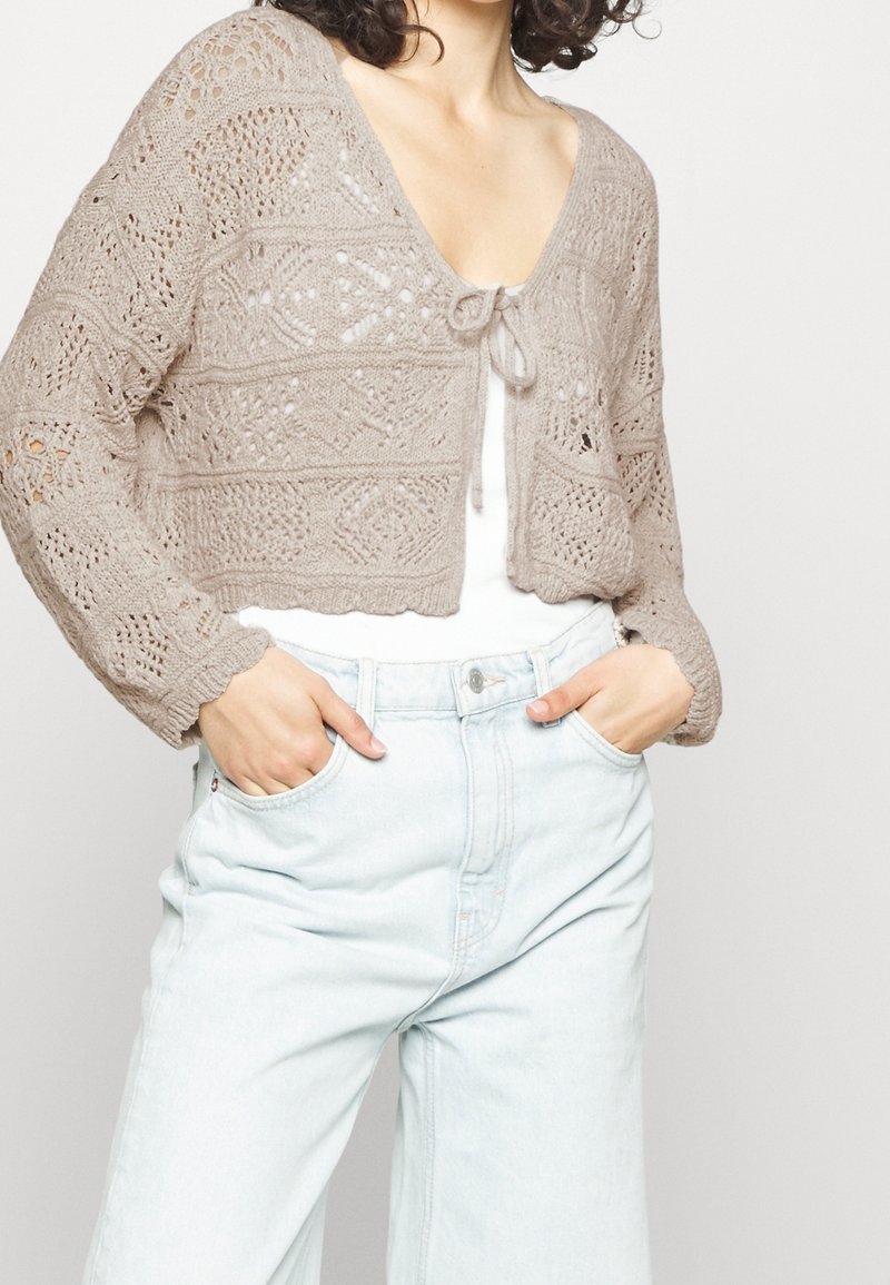 Cárdigan de punto beige con escote en V, frente abierto y patrones intrincados. Presenta mangas largas y un diseño cropped. Combinado con jeans de color azul claro.