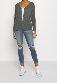 Šedý texturovaný svetr s V-výstřihem a otevřeným předním dílem, nošený přes bílé tričko. Světle modré, oprané slim džíny s roztrženými koleny a bílé tenisky.