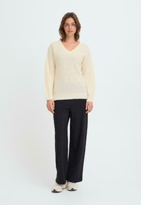 Cremfarbener V-Ausschnitt-Pullover aus strukturiertem Strick, mit langen Ärmeln und lockerer Passform, kombiniert mit schwarzen, weiten Hosen und hellen Sneakers.