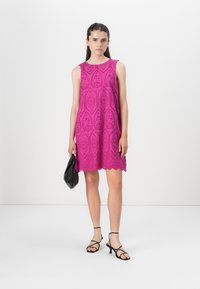 Fuksia ermeløs kjole med perforert blomsterdesign og bølget hem. Modellen holder en svart håndveske og har på seg svarte sandaler med stropper.