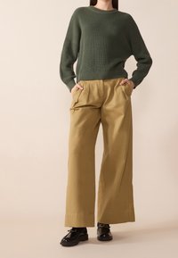 Pull vert côtelé, pantalon ample kaki avec plis, et chaussures noires à semelle épaisse. Fond neutre, mise au point sur les détails de la tenue.