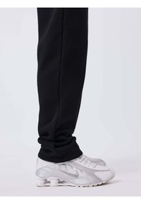 Personne portant un pantalon noir et des baskets blanches Nike avec un design de semelle visible en forme de ressort, debout sur un fond clair uni.