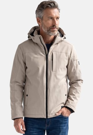 Mann mit grauem Haar und Bart, der eine beige Kapuzenjacke und blaue Jeans trägt, steht mit einer Hand in der Tasche und schaut zur Seite.