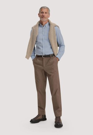 Mann mit grauen Haaren, der ein hellblaues Hemd mit Knöpfen trägt, einen beigen Strickpullover über den Schultern drapiert hat, braune Hose und dunkelbraune Schuhe.