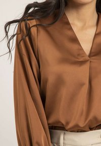 Blouse en satin marron avec un décolleté en V profond, des manches longues et une texture lisse. Conçue avec une coupe décontractée. Complétée par une ceinture de couleur claire.
