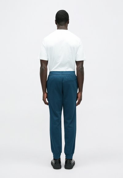 Biele tričko s krátkym rukávom a tmavomodré teplákové nohavice s elastickými manžetami, ktoré majú zadné vrecko a voľný strih. Čierne topánky dopĺňaj�ú tento vzhľad.