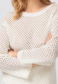 Biały dziany sweter z ażurowym wzorem o otwartym splocie i ściągaczami przy rękawach. Tekstura jest lekka i przewiewna.