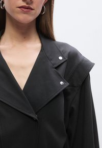 Črni plašč s strukturiranim ovratnikom, opremljen s srebrnimi pritisnimi gumbi in gladko teksturo, ki prikazuje krojen dizajn ter V-izrez na vratu.