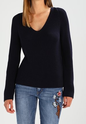 Femme portant un pull noir à col en V côtelé et un jean bleu avec une broderie florale et un tigre sur la cuisse droite.